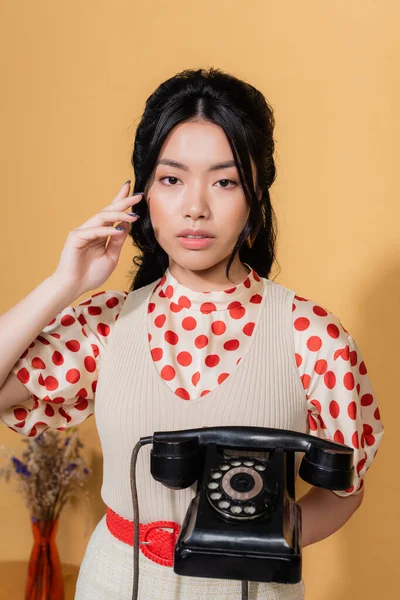 Modelo asiático sosteniendo teléfono vintage y mirando a la cámara en fondo naranja - foto de stock