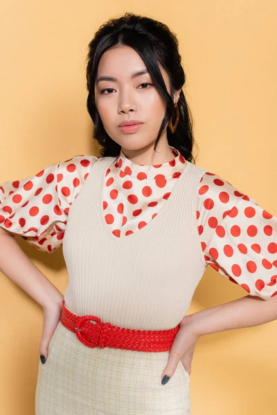 Retrato de modelo asiático elegante cogido de la mano en las caderas sobre fondo naranja - foto de stock