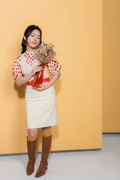 Full length of stylish asian woman in dress holding flowers on orange background — Stock Photo