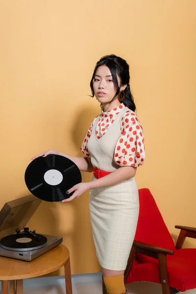 Stylish asian model looking at camera while holding record near player on orange background — Stock Photo
