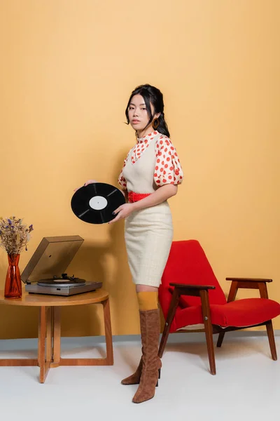 Femme asiatique en vêtements rétro tenant un record près du joueur et du fauteuil sur fond orange — Photo de stock