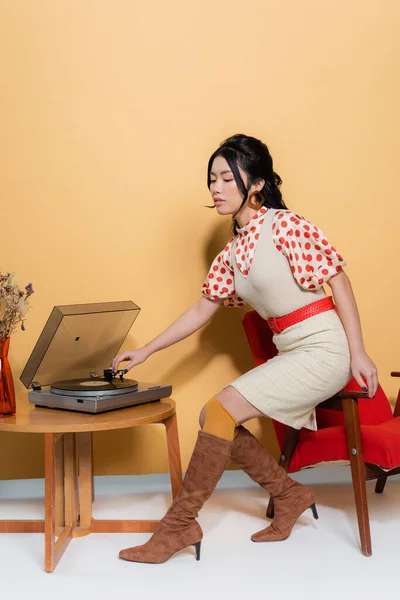 Elegante mujer asiática usando tocadiscos en sillón sobre fondo naranja - foto de stock