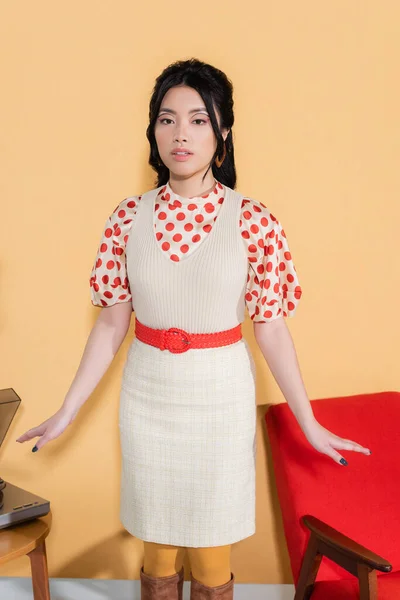 Mujer asiática de moda mirando a la cámara cerca de tocadiscos y sillón sobre fondo naranja - foto de stock