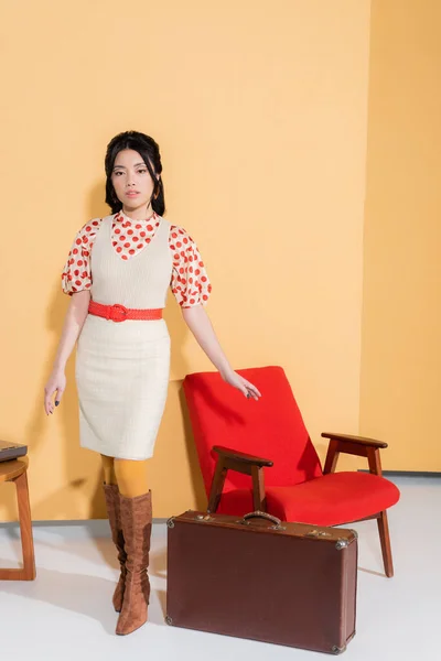 Модная азиатская модель, позирующая возле винтажного кресла и чемодана на оранжевом фоне — стоковое фото