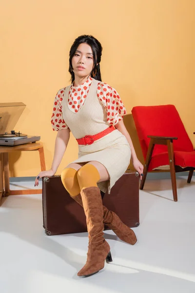 Modelo asiático de moda en vestido posando en maleta vintage sobre fondo naranja - foto de stock