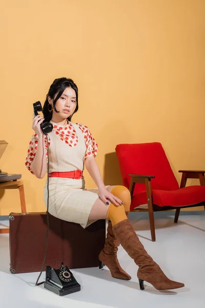 Pretty asian woman holding handset while sitting on vintage suitcase on orange background — Stock Photo