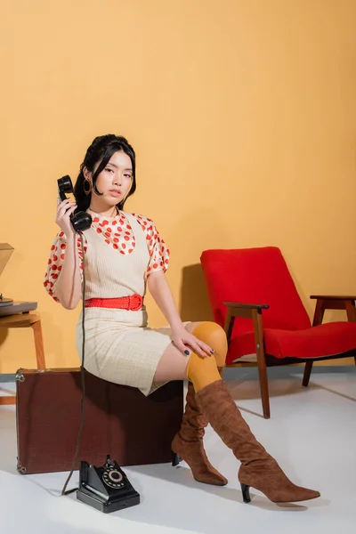 Jolie femme asiatique tenant combiné tout en étant assis sur une valise vintage sur fond orange — Photo de stock