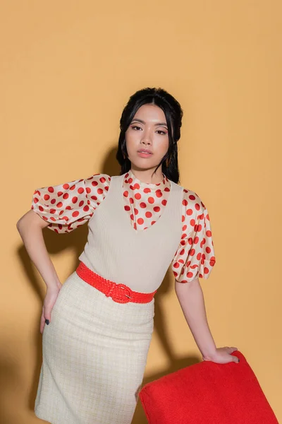 Moda asiatico donna tenendo mano su anca vicino poltrona su arancio sfondo — Foto stock