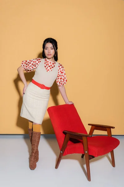 Comprimento total de mulher asiática elegante posando perto de poltrona vintage no fundo laranja — Fotografia de Stock