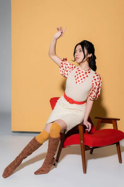 Schöne asiatische Modell in Vintage-Kleidung sitzt auf Sessel auf orangefarbenem Hintergrund — Stockfoto