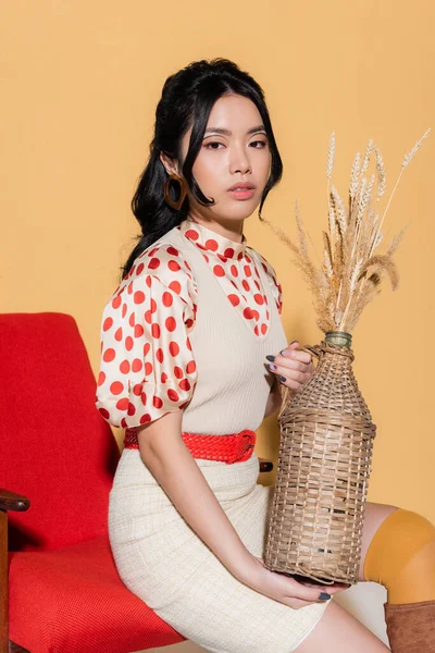 Moda asiática mulher em blusa e vestido segurando vaso com spikelets enquanto sentado em poltrona no fundo laranja — Fotografia de Stock