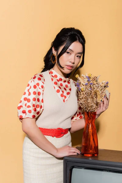 Retrato de modelo asiático con estilo en ropa vintage tocando flores en la televisión sobre fondo naranja - foto de stock