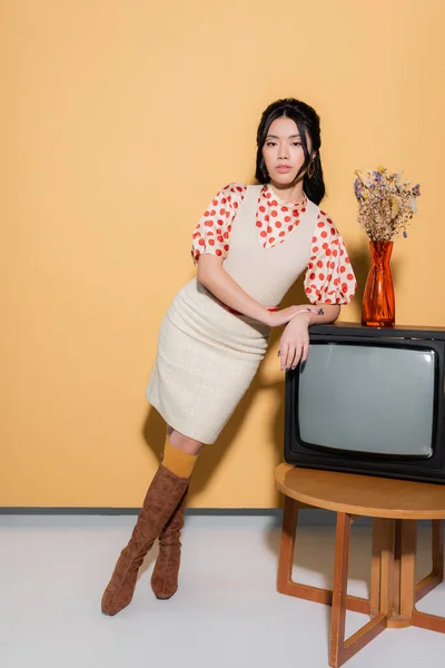 Élégante femme asiatique regardant la caméra près de la télévision rétro et des fleurs sur fond orange — Photo de stock