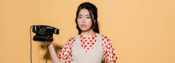 Fashionable asian model holding retro telephone and looking at camera on orange background, banner — Stock Photo