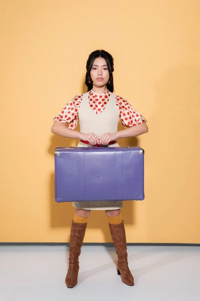 Longitud completa de elegante modelo asiático sosteniendo maleta retro sobre fondo naranja - foto de stock