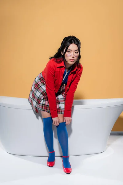 Modelo asiático ajustando calcetín de rodilla cerca de bañera sobre fondo naranja - foto de stock