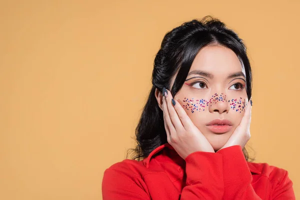 Giovane donna asiatica con brillantini sul viso guardando lontano isolato su arancione — Foto stock