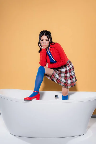 Elegante modelo asiático posando en bañera sobre fondo naranja - foto de stock