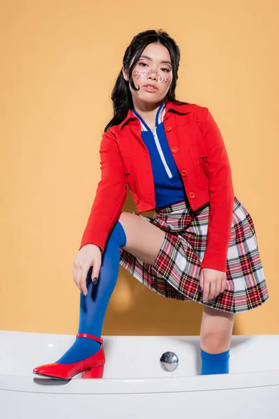 Joven mujer asiática en ropa vintage posando en bañera sobre fondo naranja - foto de stock