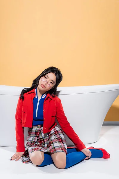 Fashionable asian model with glitter on face sitting near bathtub on orange background — Stock Photo