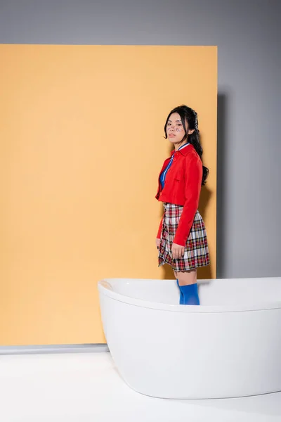Femme asiatique en vêtements rétro debout dans la baignoire sur fond orange — Photo de stock