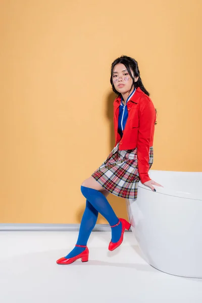 Young asian model in vintage clothes sitting on bathtub on orange background — Stock Photo