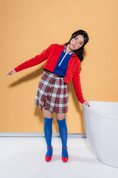 Full length of stylish asian model posing near bathtub on orange background — Stock Photo