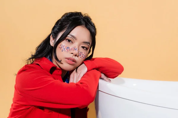 Азиатская модель с блестками на лице, смотрящая на камеру возле ванны, изолированной на оранжевом — стоковое фото