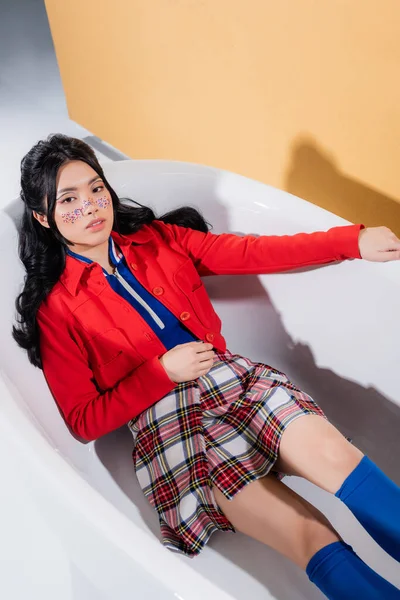 High angle view of trendy asian model lying in bathtub on orange background — Stock Photo