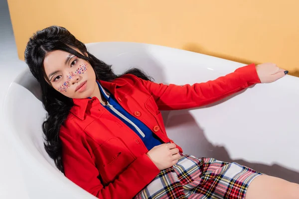 Stilvolles asiatisches Modell mit Glitzern im Gesicht, das in die Kamera in der Badewanne auf orangefarbenem Hintergrund schaut — Stockfoto