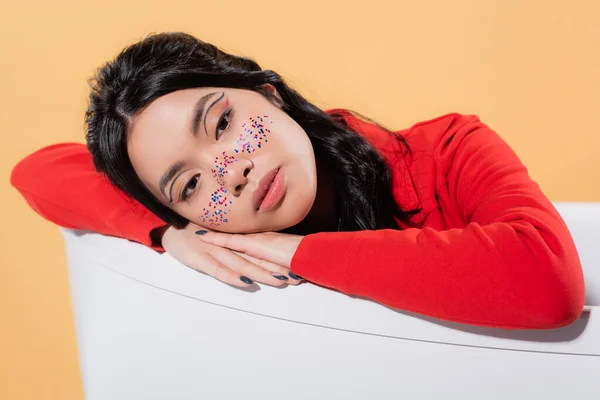 Pretty asian model with glitter on face sitting in bathtub isolated on orange — Stock Photo