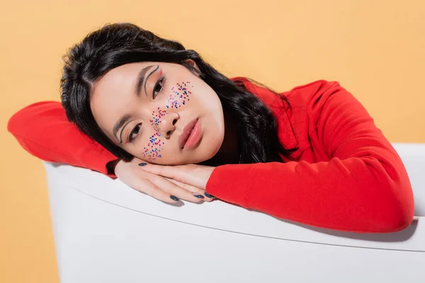 Schöne asiatische Modell mit Glitzer im Gesicht sitzt in Badewanne isoliert auf orange — Stockfoto