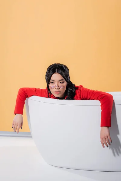 Elegante mujer asiática con maquillaje brillo sentado en la bañera sobre fondo naranja - foto de stock