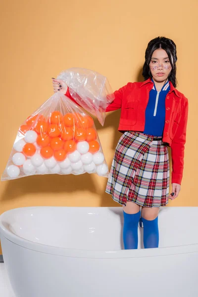 Junge asiatische Modell in Vintage-Kleidung halten Paket mit Kugeln in der Badewanne auf orangefarbenem Hintergrund — Stockfoto