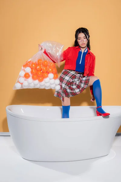 Trendy asian model in retro clothes holding package with balls in bathtub on orange background — Stock Photo