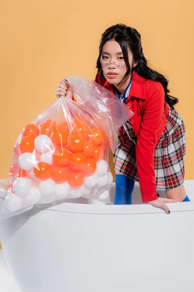Modèle asiatique à la mode avec des paillettes sur le visage tenant sac en plastique avec des boules dans la baignoire sur fond orange — Photo de stock