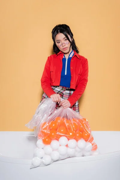 Mujer asiática de moda con maquillaje de purpurina sosteniendo bolsa de plástico con bolas en bañera sobre fondo naranja — Stock Photo