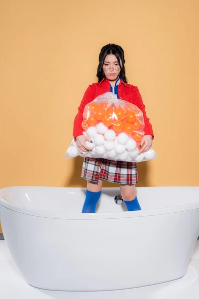 Elegante asiático mulher segurando saco de plástico com bolas e olhando para a câmera na banheira no fundo laranja — Fotografia de Stock