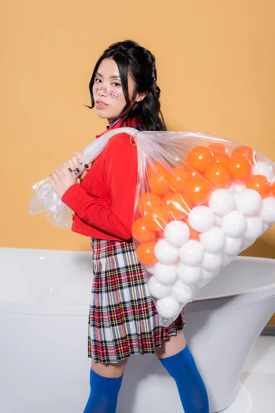 Junge asiatische Modell in Vintage-Kleidung hält Plastiktüte mit Bällen in der Nähe der Badewanne auf orangefarbenem Hintergrund — Stockfoto