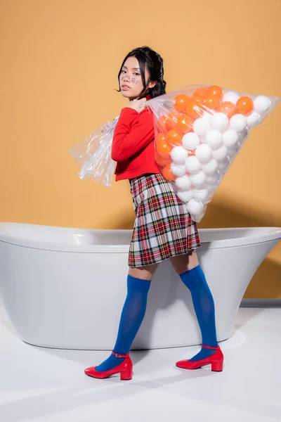 Mujer asiática de moda en ropa vintage sosteniendo bolsa de plástico con bolas cerca de la bañera sobre fondo naranja - foto de stock