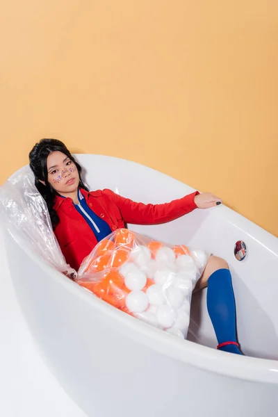 Mujer asiática de moda en ropa retro mirando a la cámara cerca de la bolsa de plástico con bolas en la bañera sobre fondo naranja - foto de stock