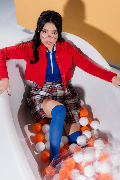 Вид зверху на азіатську жінку в ретро одязі, що сидить біля кульок у ванній на помаранчевому фоні — стокове фото