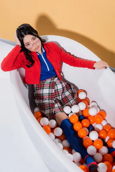 Top vista de moda modelo asiático sentado en bañera con bolas sobre fondo naranja - foto de stock