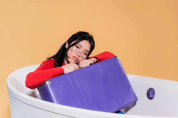 Young asian model with glitter makeup holding suitcase while sitting in bathtub isolated on orange — Stock Photo
