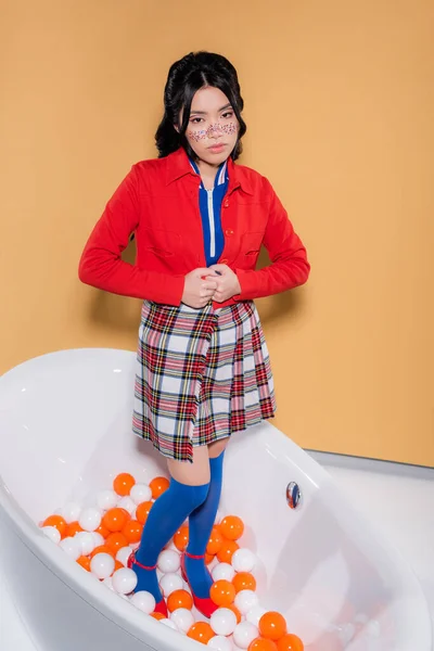 Стильна азіатська жінка дивиться на камеру, стоячи у ванній з кульками на помаранчевому фоні — стокове фото