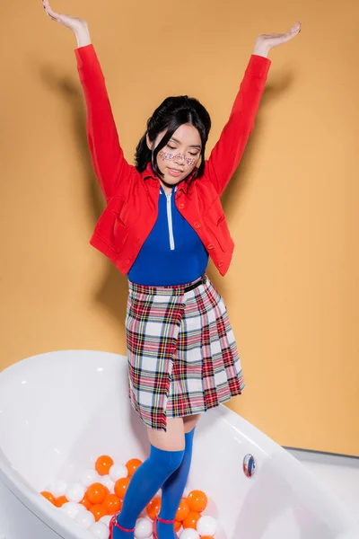 Sonriente modelo asiático en ropa retro de pie en bañera con bolas sobre fondo naranja - foto de stock