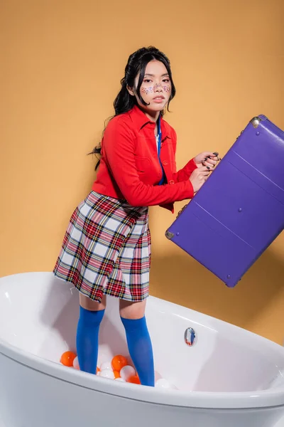 Femme asiatique à la mode avec des paillettes sur le visage tenant la valise rétro dans la baignoire avec des boules sur fond orange — Photo de stock