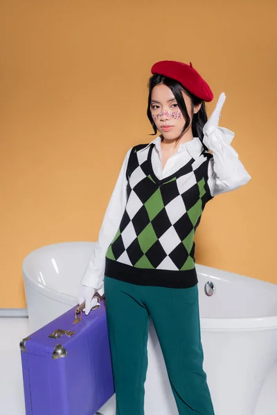 Stylish asian model in beret holding suitcase near bathtub on orange background — Stock Photo