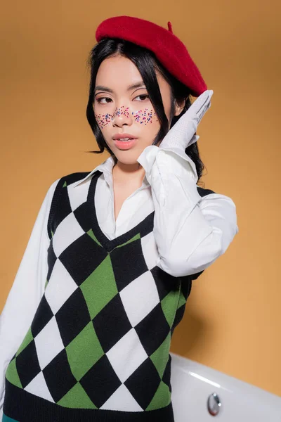 Portrait of trendy asian model in beret looking away near blurred bathtub on orange background — Stock Photo