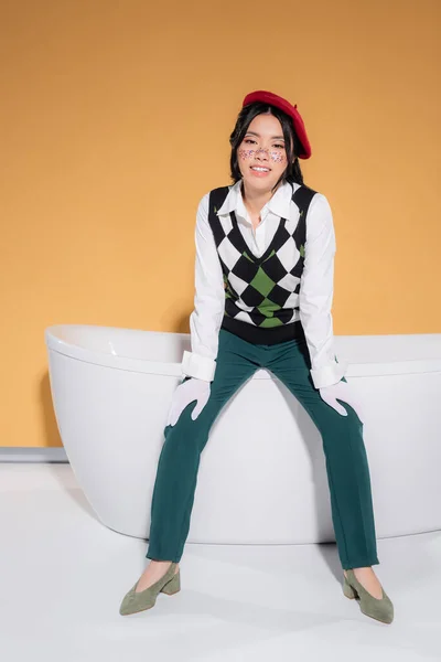 Cheerful asian model in beret sitting on bathtub and looking at camera on orange background — Stock Photo